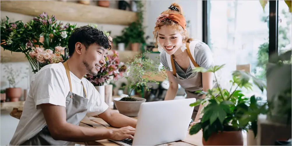 Two young people in a brightly-lit room with plants are smiling while looking at a laptop computer.