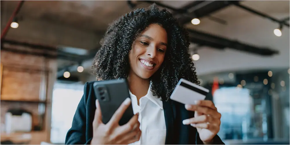 A Black woman is smiling while holding her debit card in one hand and her smartphone in the other hand.